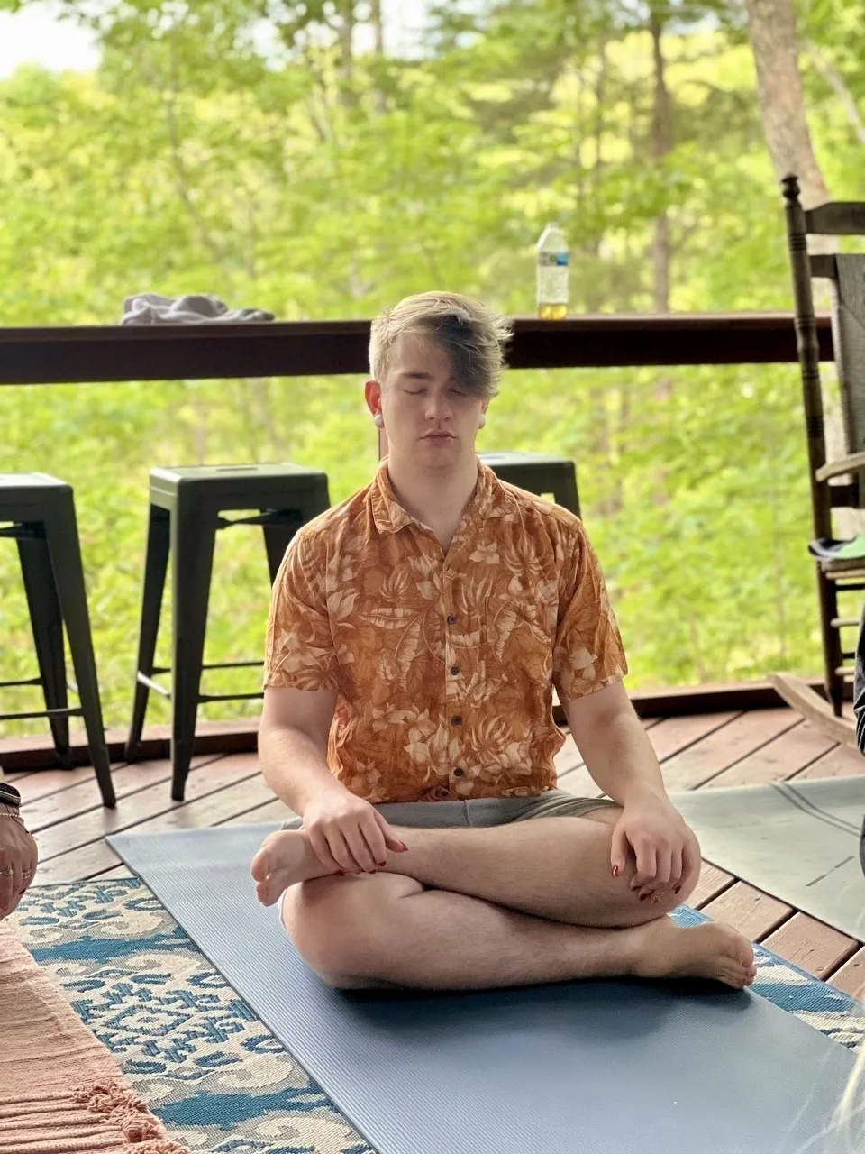 Drew seated in meditation on a porch surrounded by trees.